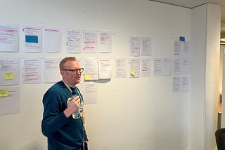A man stands in front of a while wall. He is wearing a navy blue jumper, glasses and is holding a water bottle. The wall is covered in sheets of A4 paper which are printed with information. The pages have sticky notes and pink highlighter, showing changes to the words written on them.