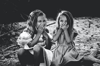 black and white image of two young girls maybe 5–6 years old, sitting on a piece of driftwood on a sandy beach. Both girls are laughing/smiling and covering their mouths with both palms. Girl on left is wearing white striped dress, girl on right is wearing grey dress. shortsleeves.