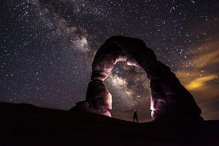 A person wearing a headlamp stands under and illuminates a stone archway in Arches National Park. The night sky and Milky Way are off to the left, and some illuminated clouds are off to the right.