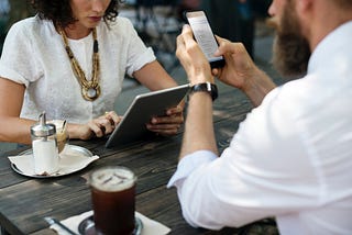 Two people sit across from each other at a wooden cafe table, one working on a laptop and the other on a cell phone.