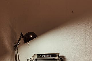 An antique typewriter is illuminated by a small black lamp agianst a white wall.