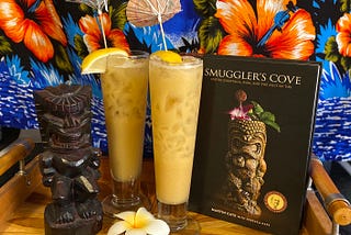 Two pale orange drinks in footed pilsener glasses on a tray next to the Smuggler’s Cove book with Hawaiian decorative items