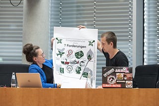 Two people in a business room setting. A woman in a blue jacket, a man with a black shirt, holding a paper with suggestions of change mobility for the better.