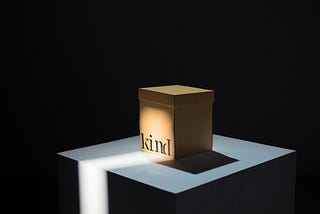 Square brown paper box sitting on a square surface. The box sits in a gleam of light and has the word ‘kind’ written on it.