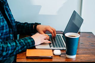 This image shows a person sitting at a wooden table, typing on a laptop.