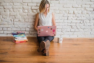 Lady sitting on a wooden floor next to a pile of books carrying a pink laptop.