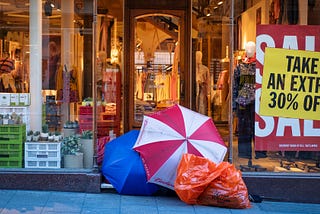 A homeless encampment made of umbrellas sits in the doorway to a high end store with sale signs in the windows.