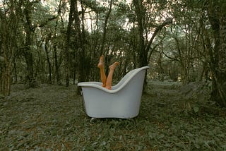 Two legs of a light-skinned person crossed over and sticking up out of a white bathtub in the middle of the woods.