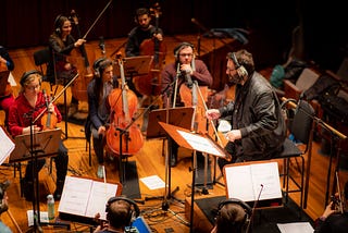 Man standing at conductor podium surrounded by orchestra, all wearing headphones with microphones recording each performer