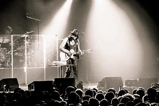 Black and white shot of a hatted Rodriguez performing with mic and guitar in front of audience, spotlight illuminates him in foreground, drummer and drum kit behind.