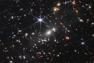 An image of hundreds of stars and galaxies seen at a distance. One bright blue star is in the center