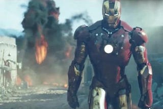 Iron Man’s gold-and-red suit, on the right side of the image, appears to be walking away from an explosion (orange flames and black smoke) in a sandy, barren area.