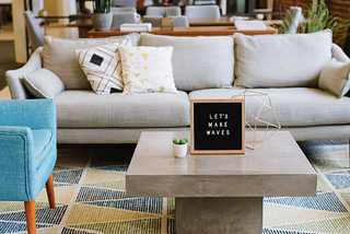 A coworking space featuring a couch, an armchair, and a concrete coffee table with a sign reading, “let’s make waves” atop it.