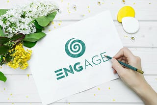 A writer’s hand writing “Engage” on a sheet of paper with green ink with flowers on the right upper corner.