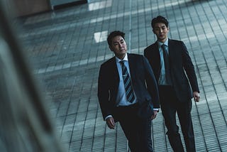 At a canted camera angle, a short, slightly pudgy east-asian man in a black suit looks into the middle-distance, walking intently next to a taller, younger east-asian man in a similarly colored suit with a slightly confused look on his face. The light is cool and blue/green. A grid of gray brick lines the floor under them, and shadows cast on it from other buildings nearby.