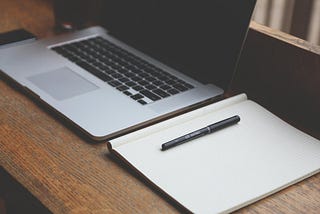 An open macbook pro with an open notebook and a pen all placed on a wood desk