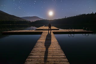 man standing at the end of a dock, surrounded by water, staring at the night sky and contemplating