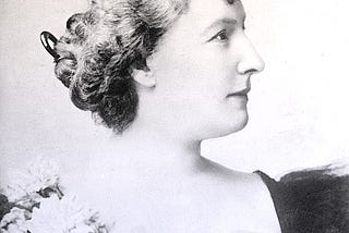 Louise in profile wearing a black ball gown and corsage. Her thick blonde hair is in a chignon.