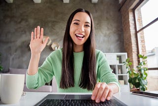 Close-up of a cheerful Asian female working from home and waving at her laptop’s webcam