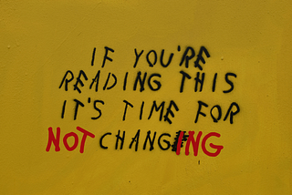 Graffiti on a yellow wall reading “If you’re reading this it’s time for change,” with the word “NOT” added and “CHANGE” altered to “CHANGING” in red paint, symbolizing resistance to unnecessary redesigns.