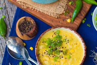 yellow corn chowder with green onions and fresh cracked black pepper, next to a blue tablecloth and peppercorn bowl