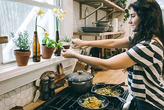 A woman in a striped shirt clips fresh basil from a potted plant above a stovetop, where noodles are prepared. The cozy kitchen features flowers, herbs, and warm natural light.