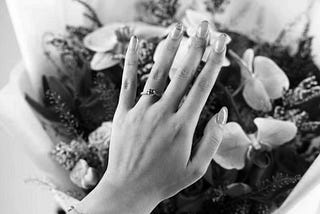 a black and white image of a woman’s hand wearing a wedding ring over a bouquet of flowers.