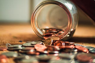 A photograph of a glass jar containing coins that has tipped over, with coins spilled on the surface in front of it.