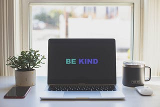 A macbook with “Be Kind” on screen. A succulent + cell phone are on the left & a yeti mug + apple mouse on the right.