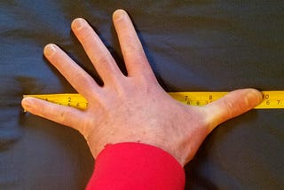 A photo of the author’s left hand with a tape measure showing a length of 9.75 inches from the tip of the pinky finger to the tip of the thumb.