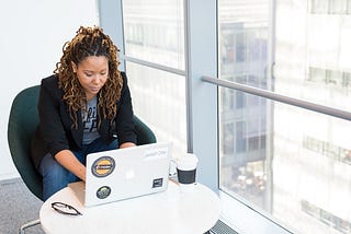 A woman sitting at a table typing on her laptop with her glasses and coffee within reach. To her left, a view of the city from the office.