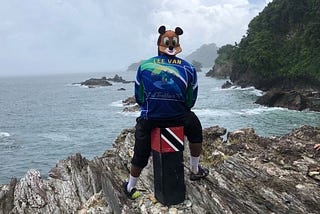 Leevan in a blue Trail Trekkers TT shirt with his name on it. His back faces us and he sits on a stool with the Trinidad and Tobago flag at the top of the stool. The stool is near the edge of a precipice and he’s facing the sea on a cloudy day.