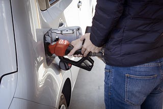 A man in a blue jack and blue jeans pumps gasoline into his white hatchback car.