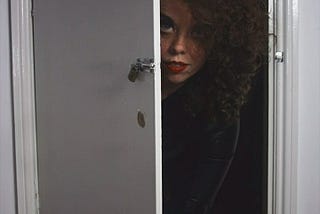 Aine, a curly haired woman is peering out of a small door at ground level. She looks as though she is crawling out and half her face is covered.