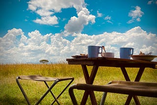 Image shows an African safari “bush breakfast” 2 camping chairs and a folding table are set in front of tall grass. On the table there are bowls and plates of varying sizes and two mugs. In the background, there is a bright blue sky dotted with white, fluffy clouds