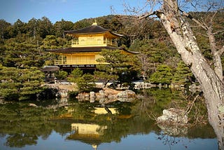 Kinkaku-ji (Golden Pavilion) in Kyoto, Japan, a three-story Zen Buddhist temple with gold-leaf-covered upper floors, reflected in a tranquil pond and surrounded by lush greenery. A large tree trunk appears in the foreground, adding depth to the serene landscape.