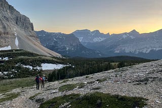 Two hikers on a rocky trail with mountains and a sunset in the background.