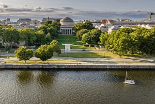 Aerial photo of MIT’s Great Dome with a lawn, trees, and a river in the foreground.