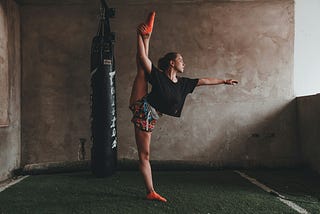 A woman in a basement by a punching bag stretches her leg above her head