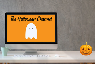 White desk in front of gray background. Computer monitor on desk shows The Halloween Channel title and logo ghost on orange background. Jack ‘O Lantern sits on desk.