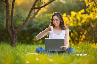 Woman working on a laptop sitting in a green field thinking