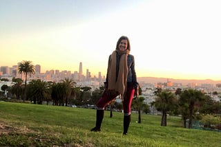 The author, a young woman, stands in a tree-lined park smiling. The San Francisco, California skyline and a sunset fill the background. She’s wearing a tan scarf, jean jacket, and pink yoga pants.