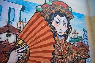 image features a colorful mural painting on a wall, depicting a stylized female figure dressed in traditional Chinese attire. The woman has an intense gaze, pale skin, and dark, wavy hair adorned with an ornate red and gold headdress decorated with green gemstones. She holds a large, red fan with sunburst-like stripes in her right hand. The mural also includes architectural elements resembling pagodas and classical Chinese buildings in the background, along with a blue sky and abstract structure