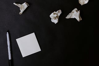 A white stack of sticky notes sits on a black surface. To the left is an un-capped pen waiting to be used. Also on the surface are four crumpled balls of discarded ideas.