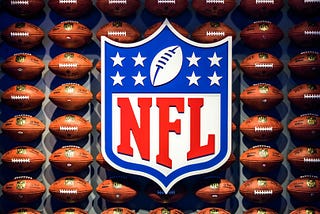 An official red, white, and blue, official NFL badge in front of 8 rows of footballs lined up against a wall. The NFL badge has the NFL logo, 8 stars, and a football on top.