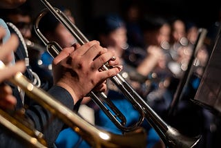 A picture of an orchestral brass section. The photo zooms in on a trumpet player’s hands. In the blurred background, you can see the french horn section.