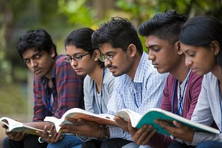 This image shows a group of Indian students sitting outdoors, each reading a book. They appear focused and engaged in a group study session, with a natural, green background adding to the calm atmosphere.