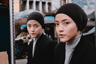 Woman with black head covering and jacket, and grey jersey top covering her neck, standing before a mirror which also shows her reflection. The reflection looks directly at the viewer which is incongruent with the woman outside her reflection, suggesting editing tricks/artwork to give this effect. Background is a street possibly in East Asia or China with an open air stall in the mirror reflection.