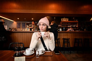 white woman with short brown hair, pink knitted cap, white shirt, holding small porcelain coffee cup in a café, looking away from the camera
