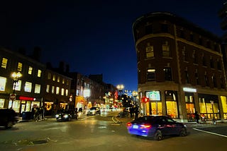 Street scene at night. Non-descript buildings with their lights on and cars passing on the street, a few pedestrians are walking on sidewalks.